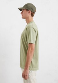 Olive green cotton t-shirt with short sleeves and a crew neckline, worn with light-colored pants and a matching baseball cap.