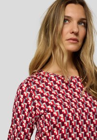 Rotes und cremefarbenes Oberteil mit Rundhalsausschnitt, ausgestattet mit geometrischen Formen in verschiedenen Schattierungen, hergestellt aus einem weichen Stoffgemisch.