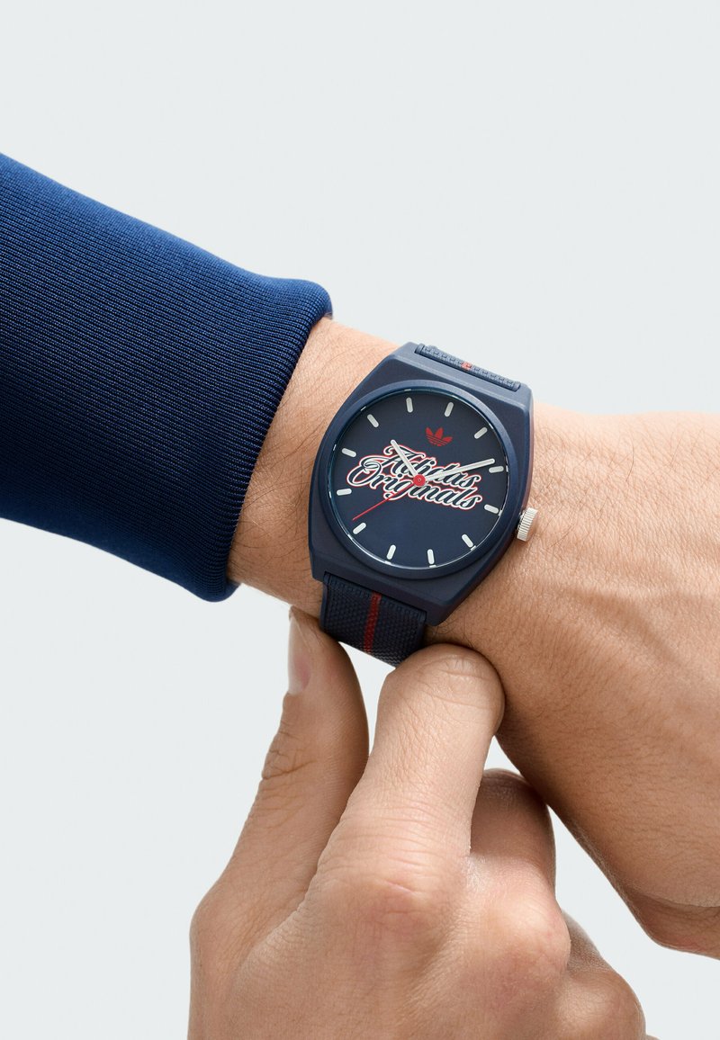 Person passt dunkelblaue Adidas Originals Armbanduhr mit marineblauem Armband und rotem Streifen am linken Handgelenk an, trägt ein blaues Langarmshirt.