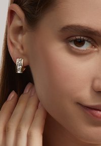 Goldfarbene Creolen mit rechteckigen klaren Steinen, glatter Textur und modernem Design. Hand berührt sanft das Ohrläppchen.