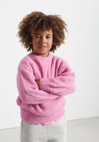 Ružový pletený sveter s textúrovaným vzorom, okrúhlym výstrihom a dlhými rukávmi, doplnený o šedé tepláky. Modelka stojí s prekríženými rukami.