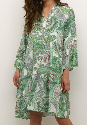 Vrouw draagt een losse, lange mouw, knie-lange jurk met een groen, wit en roze paisley patroon en een gedeeltelijke knoopsluiting bij de kraag.