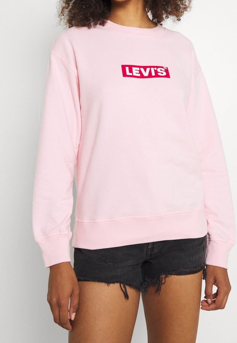 Personne portant un sweat Levi’s rose clair et un short en jean noir effiloché, debout devant un fond blanc uni.