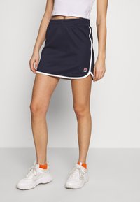 Jupe de sport bleu marine avec bordure blanche, taille élastique et logo Fila ; portée avec des baskets blanches et des chaussettes chevilles orange.