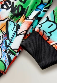 Kolorowy sweter z nadrukiem w stylu graffiti, przeważnie w zielonym, pomarańczowym i niebieskim kolorze. Czarnym, prążkowane mankiety dodają tekstury.