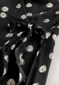 Rochie neagră cu un model pătrățel și desene circulare albe, țesătură texturată și detaliu la talie legat. Prezintă un decolteu în V și o siluetă fluida.