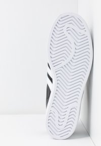 Mustat lenkkarit valkoisella pohjalla, joissa on teksturoitu kumipohja ja kolme valkoista vinoa raitaa sivussa.