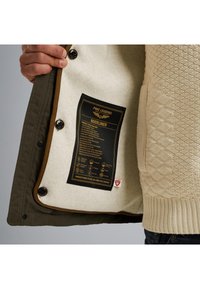 Maslinasto zeleni kaput s bež unutarnjosti, koji ima teksturirani pleteni detalj i crnu etiketu za njegu s uputama za njegu.