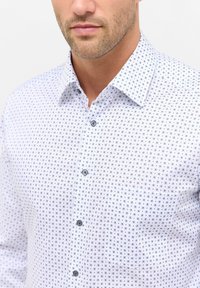 Camicia button-up in tessuto bianco con motivi circolari blu, dotata di colletto aperto e bottoni grigi; texture liscia, design slim fit.