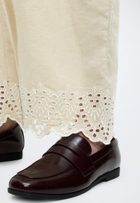 Σκούρα καφέ δερμάτινα μοκασίνια φορεμένα με μπεζ παντελόνι που έχει λευκές κεντητές λεπτομέρειες με κοψίματα στο τελείωμα.