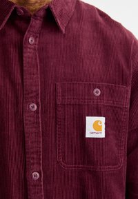 Bordeaux kordfløjlsskjorte med knapkrave, der har en enkelt brystlomme og et hvidt mærke med et orange logo. Knapperne passer til skjorten.
