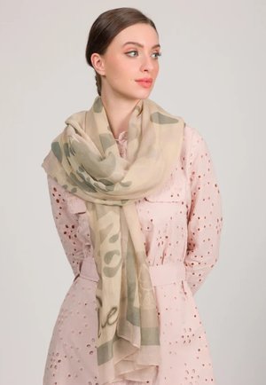 Giovane donna che indossa un vestito rosa chiaro con motivi a occhiello e una sciarpa beige decorata, che guarda verso destra su uno sfondo uniforme.