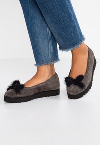 Zapatos planos de ante gris con un acento de piel de pelo negra en la punta y una suela de goma negra gruesa. Se llevan unos vaqueros con dobladillos desgastados con los zapatos.