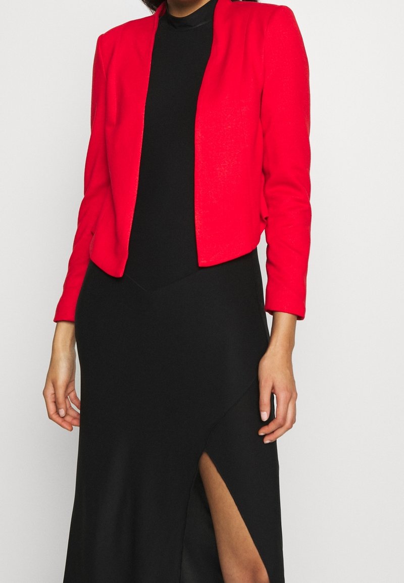 Kvinna iklädd en klar röd kavaj över en svart klänning med en hög skårad slits, står framför en enkel vit bakgrund.