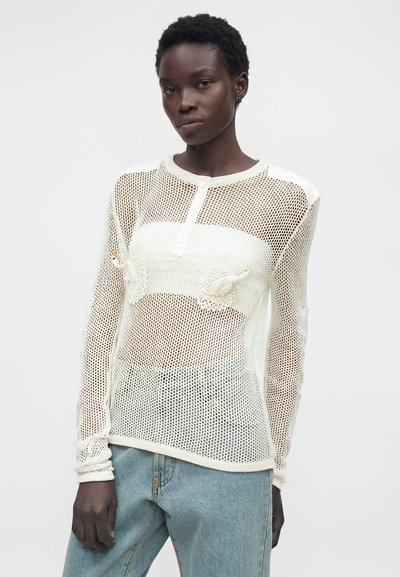 Femme portant un haut henley à manches longues en maille blanche sur un bandeau blanc, associé à un jean bleu clair, debout devant un fond uni.