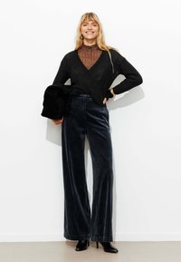 Pull en tricot noir avec un col en V, associé à un pantalon en velours côtelé large bleu foncé. Le mannequin tient un sac noir et porte des chaussures à talons hauts noires.