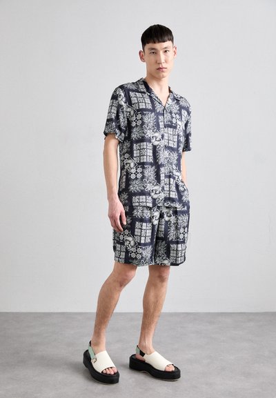 Short-sleeve navy shirt and matching shorts featuring white floral and geometric patterns. Worn with off-white sandals and a thick black sole.