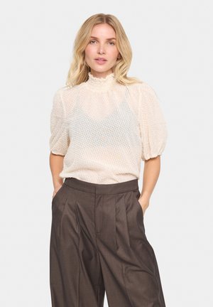 Blusa di tessuto crema con maniche a palloncino e colletto alto, abbinata a pantaloni plissettati larghi di colore marrone scuro. Il tessuto ha una leggera lucentezza.
