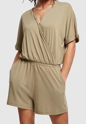 Kvinde iført beige kortærmet romper med slå-om front, elastisk talje og sidelommer, accessoriseret med lagdelte guldkæder.