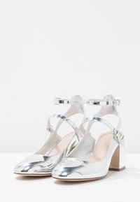 Scarpe con tacco metallico argento, con schienale aperto, cinturino alla caviglia e punta arrotondata. Tacco a blocco e superficie liscia e riflettente.