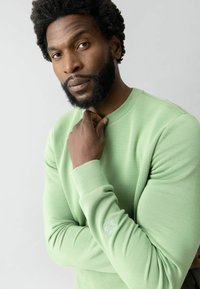 Hombre con barba y cabello rizado, vestido con una camisa de manga larga verde claro, tocándose el cuello, mirando pensativo a la cámara sobre un fondo claro.