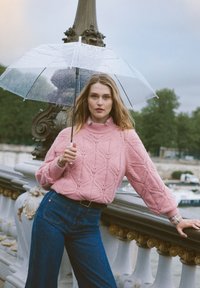 Roosa kõrge kaelusega kampsun koos mustrilise tekstuuriga, kombineeritud siniste laiade säärega teksadega. Selge vihmavari kaitseb vihma eest.