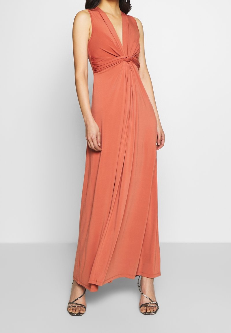 Robe maxi corail sans manches avec un détail de nœud torsadé à la taille, encolure en V, portée avec des sandales à talons hauts à brides fines.