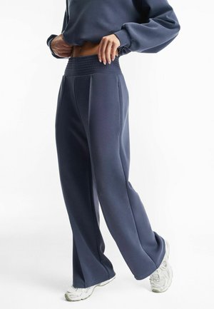 Osoba ubrana w szerokie, granatowe spodnie dresowe i pasującą bluzę, poprawiająca pas, a na stopach ma widoczne białe buty sportowe.