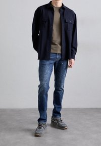 Námořnická košile na knoflíky přes olivové tričko, v kombinaci s modrými džíny se sprejovými skvrnami. Šedé tenisky celý outfit doplňují.