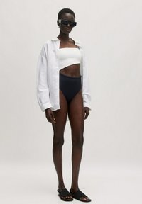 Witte oversized shirt, zwarte hoge bikini, en zwarte sandalen. Model draagt grote vierkante zonnebril. Glad materiaal en een aansluitend ontwerp.