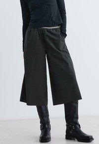 Person in weiten, knöchellangen schwarzen Hosen, schwarzem langärmeligen Oberteil und schwarzen Lederstiefeln mit Schnallen, steht vor einem schlichten hellen Hintergrund.