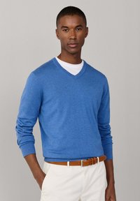 Homme portant un pull bleu à col en V par-dessus une chemise blanche, pantalon crème avec ceinture marron, debout devant un fond gris uni.