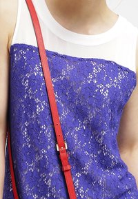 Top in pizzo blu con scollatura bianca, caratterizzato da un motivo floreale. Una sottile striscia rossa attraversa il design, arricchendo l'aspetto a strati.