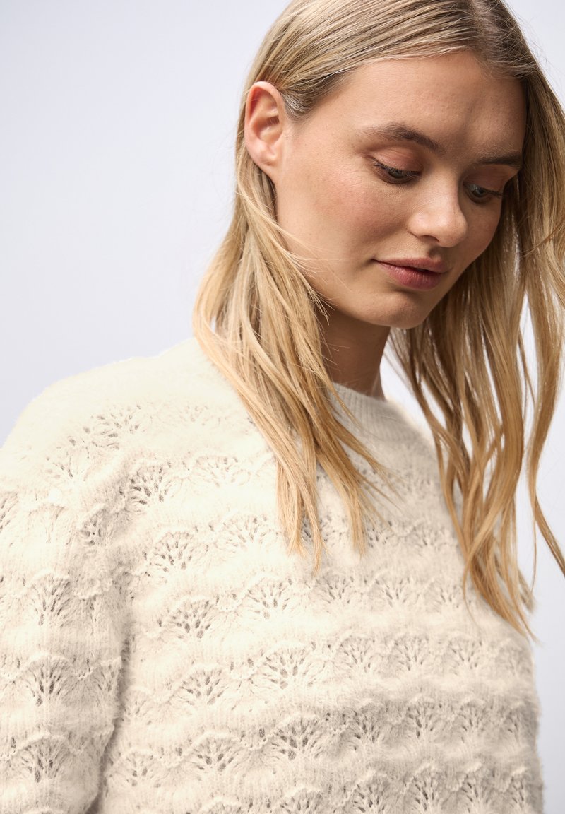 Suéter de punto color crema con un diseño de patrón de olas. Textura suave, escote redondo y mangas largas, parcialmente cubierto por el cabello.