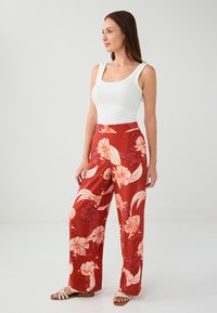Brede broekspijpen in een rode stof met grote witte bloemenpatronen, gecombineerd met een aansluitend wit tanktop en lichte sandalen.