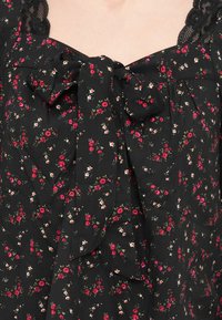 Μαύρο ύφασμα με φλοράλ μοτίβο σε κόκκινες και λευκές αποχρώσεις, με φιόγκο στον λαιμό και δαντελένιες λεπτομέρειες στους ώμους και τον λαιμό.