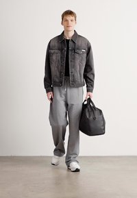 Model wearing a black denim jacket over a black T-shirt, gray sweatpants, and white sneakers, holding a black leather bag.