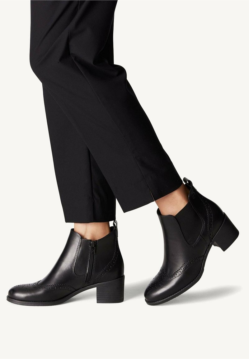 Personne portant des bottines noires à talons blocs bas et détails brogues, associées à un pantalon noir raccourci sur un fond uni.