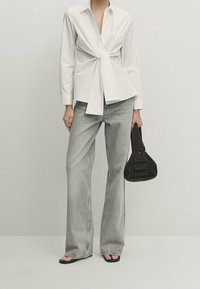 Hvid skjorte med knappelukning og frontknude, parret med lysegrå brede jeans og en mørkebrun struktureret håndtaske. Sorte slip-on sandaler.