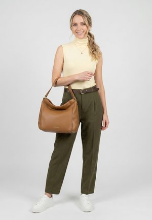 Mujer sonriente con cabello ondulado lleva una blusa sin mangas amarilla, pantalones oliva, zapatillas blancas y sostiene un bolso de cuero marrón por la correa.