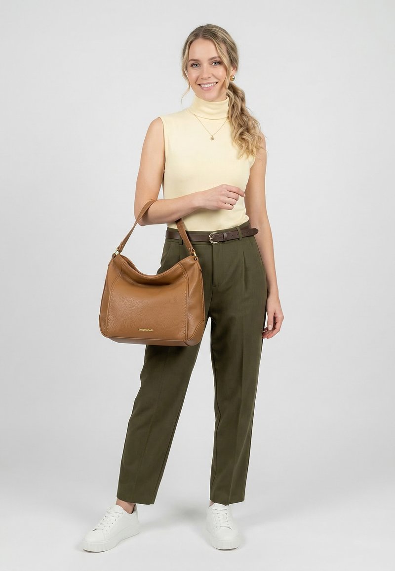 Femme souriante aux cheveux ondulés porte un haut jaune sans manches, un pantalon olive, des baskets blanches, et tient un sac à main en cuir marron par sa anse.