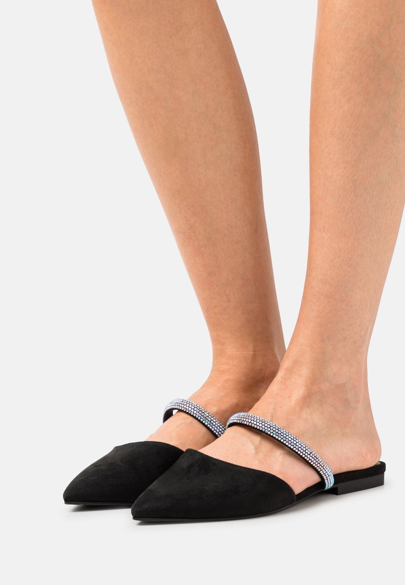 Paire de ballerines noires à bout pointu, à enfiler, avec des fines brides scintillantes, portées pieds nus sur un fond clair uni.
