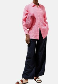 Camicia a righe rosa con bottoni e maniche lunghe, abbinata a pantaloni larghi scuri e sandali con slip-on a fantasia. Tiene una borsa rosa testurizzata.