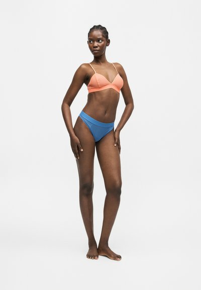 Γυναίκα που στέκεται ξυπόλητη, φοράει ένα ροδακινί σουτιέν με λεπτές τιράντες και μπλε σλιπ με ψηλή μέση, κοιτάζει ελαφρώς προς τα αριστερά, σε απλό λευκό φόντο.