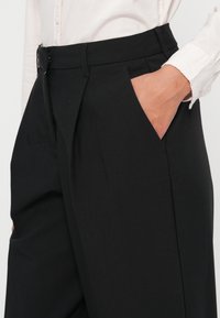 Black trousers featuring a smooth texture, pleated front, and side pockets, paired with a white button-up shirt.