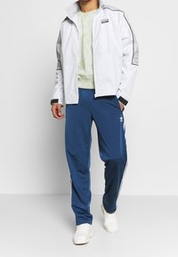 Bílá zipová bunda s šedými akcenty, zelená mikina s tie-dye vzorem a modré tepláky s bílými pruhy. Nesené s bílými teniskami.