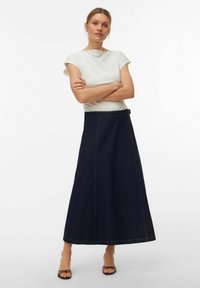 Jupe maxi en denim bleu foncé avec une silhouette droite, des coutures jaunes et une fermeture latérale. Associée à un haut blanc ajusté.