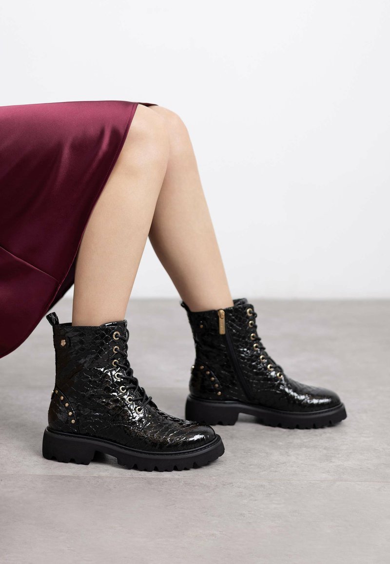Botas negras y brillantes con un patrón de reptil texturizado, con cordones en la parte frontal y cremallera lateral. Suela de goma negra resistente con tachuelas decorativas.