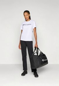 Mulher vestindo uma t-shirt branca da Champion e calças pretas, segurando uma mala de viagem preta da Champion, diante de um fundo claro e liso.