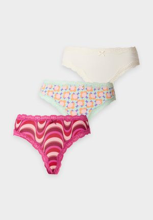 Frau mit langen dunklen Haaren, die einen schwarzen BH und pink gemusterten Slip trägt, steht barfuß vor einem schlichten weißen Hintergrund.